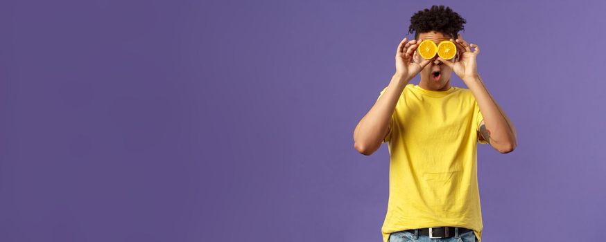 Holidays, vitamins and vacation concept. Portrait of funny, playful young guy fool around, playing with fruits, making eyes from pieces of orange, show wondered face expression, purple background.