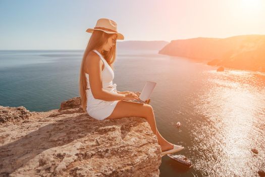 Successful business woman in yellow hat working on laptop by the sea. Pretty lady typing on computer at summer day outdoors. Freelance, travel and holidays concept.