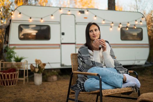 Beautiful caucasian woman is sitting wrapped in a plaid and drinking a warming drink outdoors. Traveling in a motor home in autumn