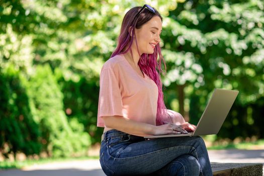 Young girl with pink hair is studying in the spring park, sitting on the wooden bench and browsing on her laptop