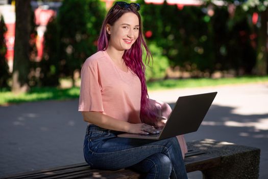 Young girl with pink hair is studying in the spring park, sitting on the wooden bench and browsing on her laptop