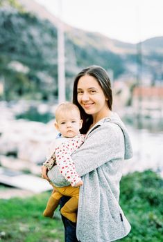 Mom with a baby in her arms stands on the shore of the marina against the background of the masts of boats. High quality photo