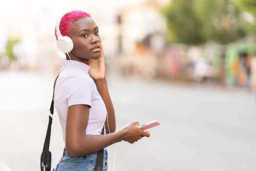 Afro young woman with short pink hair listening to music using the mobile in the street