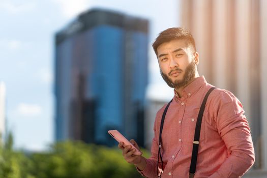 Asian businessman in a shirt and braces looking at the camera while using a mobile outdoors