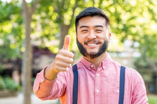 Smiling an asian man in casual clothes gesturing success with the hand in a park