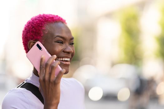 Stylish afro young woman with short pink hair smiling while talking to the mobile in the street