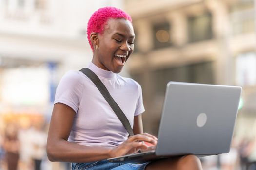 Stylish young woman laughing while using a laptop on the street