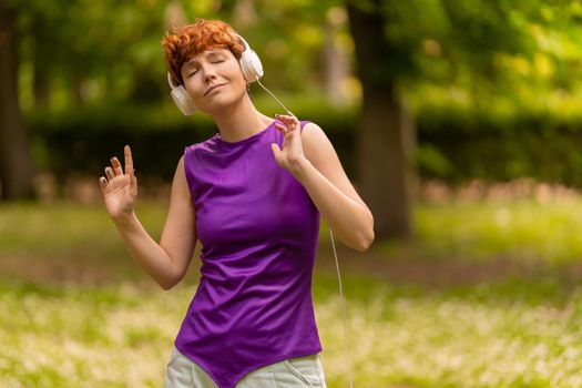 Relaxed non binary meloman in casual clothes with short ginger hair closing eyes and dancing while listening to music on weekend day in park