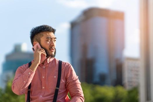 Asian man in a shirt and braces calling on the phone with skyscrapers in the background