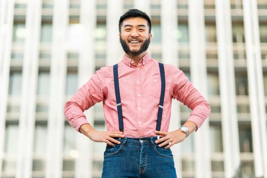 Smiling asian man in casual clothes posing with his arms on his hips looking at the camera outdoors