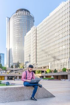 Vertical photo of a asian man in a shirt and braces working on a laptop in a square in a financial district