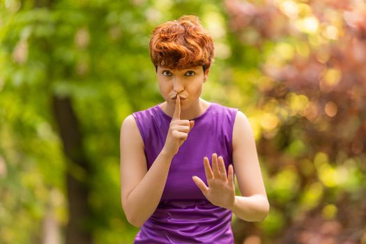 Redhead female of non binary gender identity making shh gesture and looking at camera while spending time in lush park