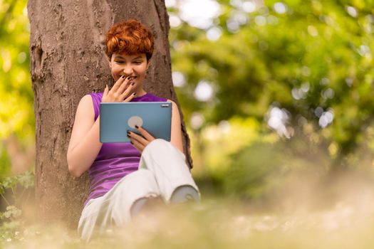 Happy non binary person in casual clothes with short ginger hair covering mouth and giggling, while sitting near tree trunk and watching funny video on tablet on sunny weekend day in park