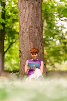 Female of non binary gender identity leaning on tree trunk and watching video on tablet while sitting on lawn on summer weekend day in park