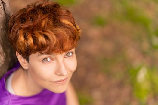 From above of positive lesbian female with short red hair smiling and looking at camera while resting near tree in park in summer