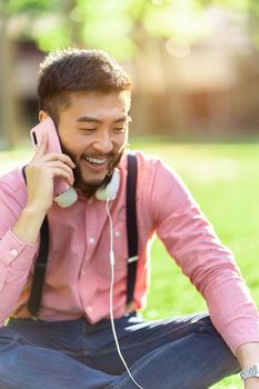 Asian man in casual clothes with headphones in his neck laughing and talking on his mobile in a park