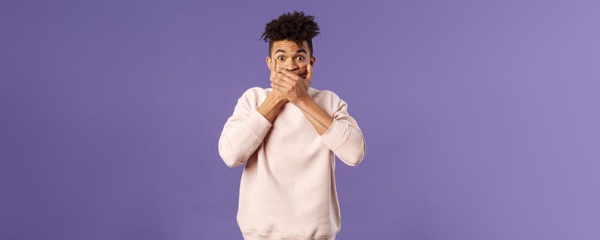 Portrait of shocked impressed young hispanic man found out gossip, heard rumour, trying not laugh shut his mouth with hands and express amazement and surprised with gaze, purple background.