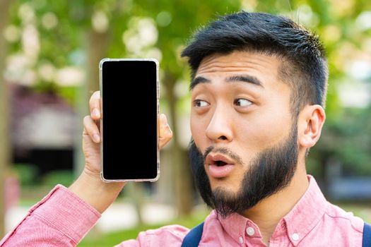 Portrait of a chinese man with a surprised face showing the screen of a mobile phone in a park