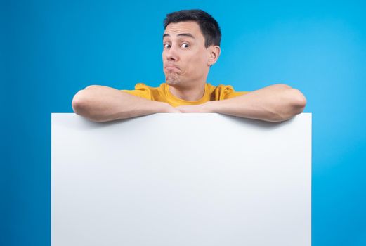 Excited male raising eyebrows and widening eyes while offering empty mockup space and staring at camera against blue background