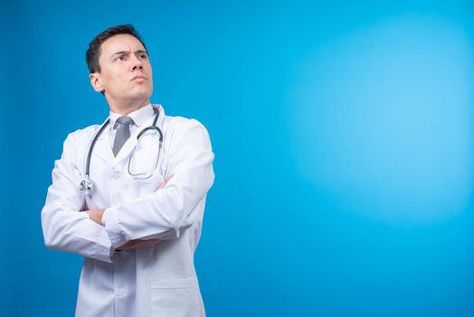 Isolated male physician in white uniform crossing arms and looking away bravely while standing on blue background