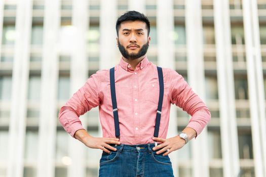 Serious asian man in casual clothes posing with his arms on his hips looking at the camera outdoors