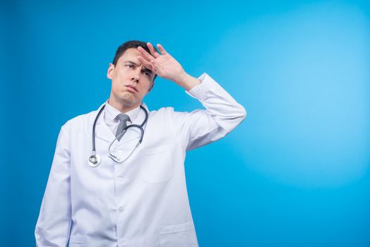 Physician with stethoscope touching forehead and looking away while being tired at work on blue backdrop