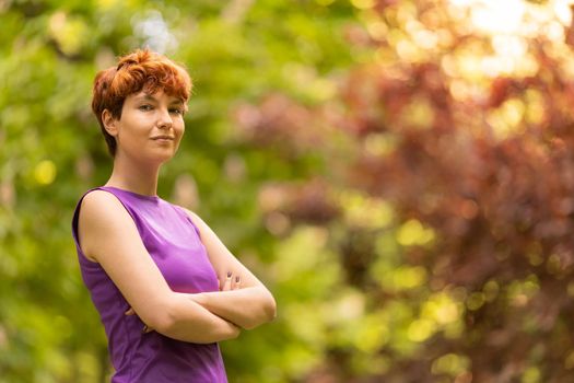 Confident non binary person in purple top with short ginger hair crossing arms and looking at camera, while standing on blurred background of trees on sunny day in park