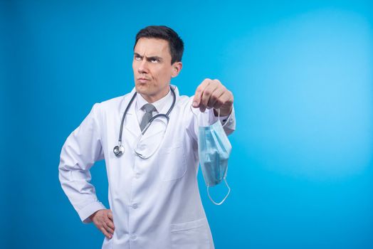 Displeased doctor in medical uniform with stethoscope looking away on blue background while holding protective mask with hand on waist