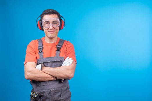 Positive male carpenter in uniform with protective goggles and earmuffs crossing arms and looking at camera with smile against blue background