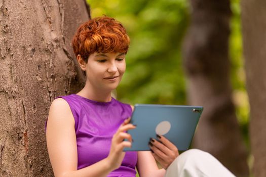 Redhead androgynous female in purple top with shot red hair sitting near tree trunk, and watching video on tablet while spending time on weekend day in summer garden