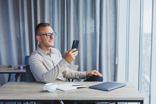 A young attractive male freelancer in glasses sits in the office and looks at the phone. Handsome blogger writing ideas on phone while sitting at wooden table in office with laptop