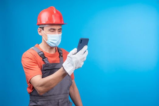 Man in workwear with gloves, face mask and hardhat watching video on mobile phone while taking break in work against blue background during pandemic