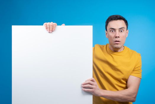 Surprised young male in yellow t shirt looking at camera and demonstrating blank sheet of paper against blue backdrop