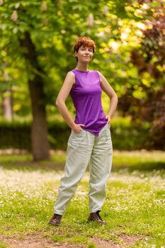 Full body of positive androgynous female in casual clothes with short red hair holding hands in pockets, and looking at camera on blurred background of green trees in summer park