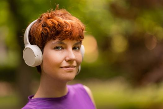 Glad androgynous redhead woman listening to music in white headphones and looking at camera on blurred background of park in summer