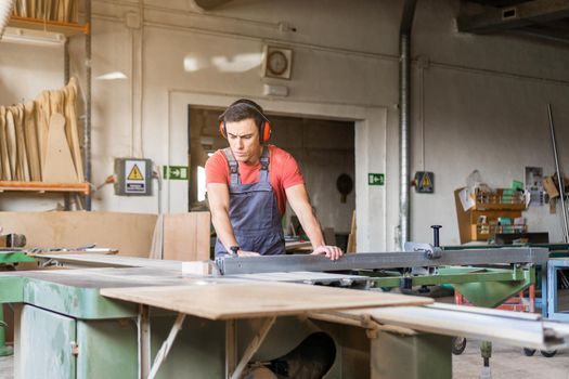 Focused male woodworker in uniform cutting wooden plank with squaring machine while working in light spacious carpentry with various equipment