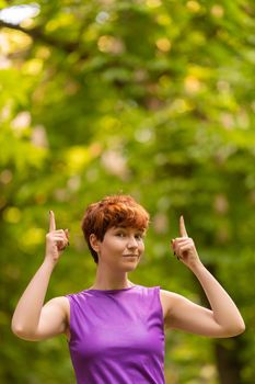 Positive non binary female in purple top with short ginger hair looking at camera and pointing up against lush trees on summer day in park