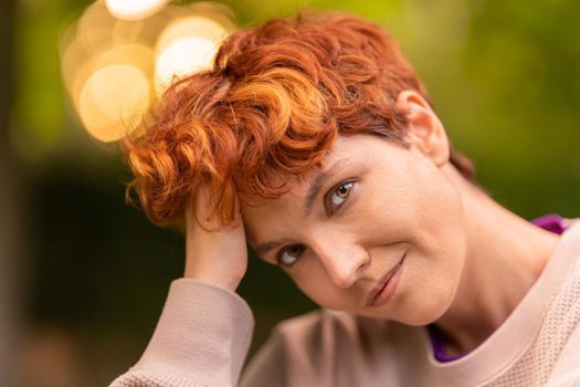 Glad androgynous woman touching short red hair and looking at camera while spending summer day in park