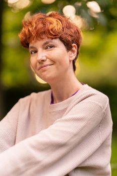 Glad lesbian female in casual sweatshirt with short ginger hair smiling and looking at camera on blurred background of summer park