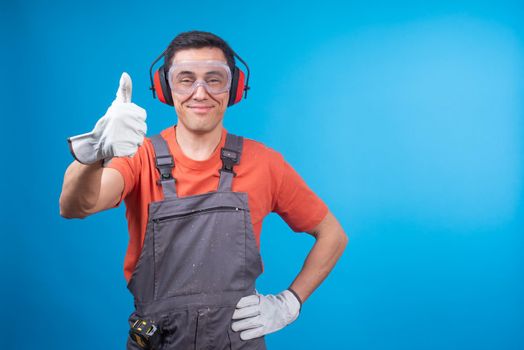 Satisfied craftsman in workwear with gloves, protective goggles and earmuffs looking at camera with smile, holding hand on waist and gesturing thumb up while approving work against blue background