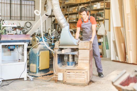 Focused male worker in uniform working on professional thicknesser machine while trimming wooden plank in light joinery with professional equipment