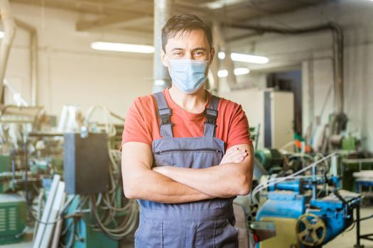 Professional male carpenter in protective mask looking at camera with crossed arms while standing in light carpentry with various equipment