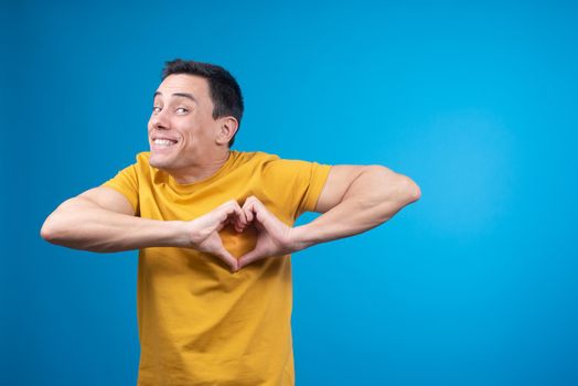 Cheerful young male model smiling brightly and showing gesture of romantic love and looking at camera on blue background
