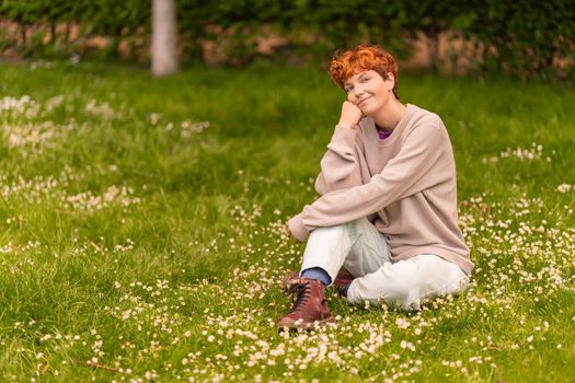 Positive androgynous female in casual outfit leaning on hand and looking at camera, while resting on grassy meadow with white flowers on summer day in park