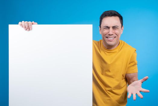 Bewildered male model showing empty white poster and looking at camera with frowning face from confusion and misunderstanding against blue background