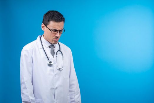 Isolated dissatisfied physician in glasses and medical coat with stethoscope looking down on blue background