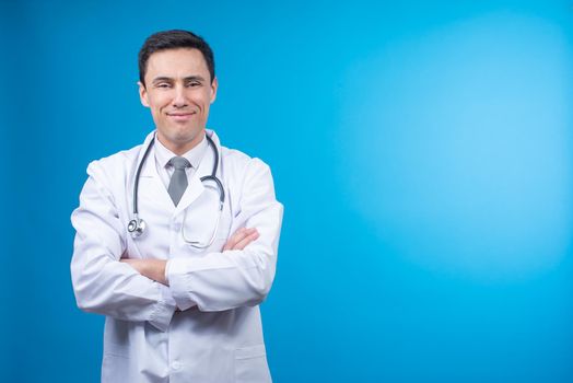 Pleasant doctor in medical uniform standing on blue background and looking at camera delightfully with stethoscope on neck