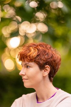 Redhead androgynous woman with short hair looking away on blurred background of park in summer