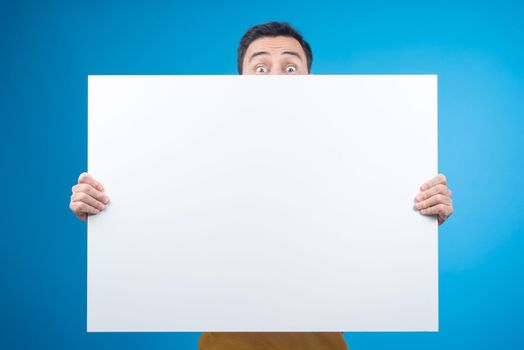 Astonished male model covering face with blank banner and staring at camera with eyes wide open on blue background