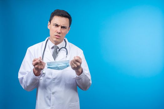 Physician in white robe with stethoscope frowning and looking at camera on blue background while holding mask